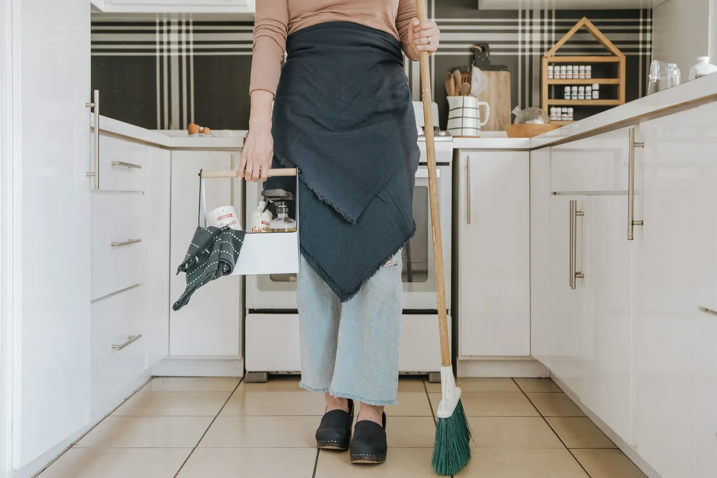A cleaner in an apron holding a broom in a bright, tidy kitchen
